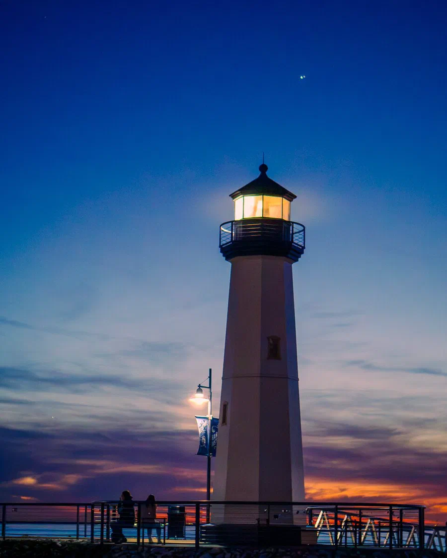 El faro de Rockwall Harbor iluminado al anochecer con el cielo azul y naranja; arriba a la derecha brillan juntos Júpiter y Saturno