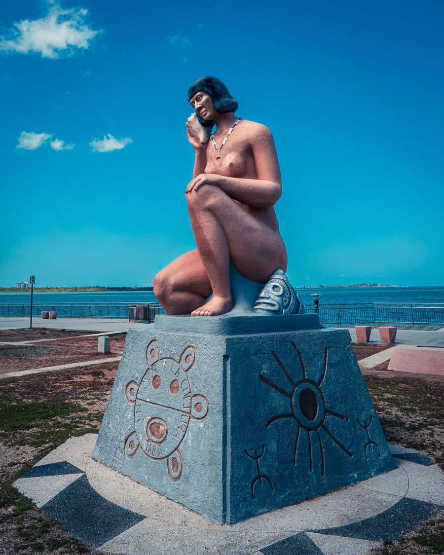 Estatua de una mujer taína sentada que se lleva un caracol al oído, junto a una cara de piedra, sobre una base azul con un sol y figuras taínas talladas, frente al agua bajo un cielo azul, en Cataño, Puerto Rico