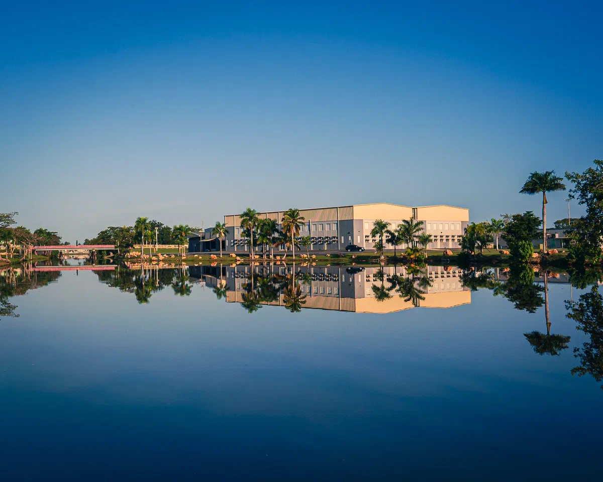 Un edificio claro entre palmas se refleja completo en el agua quieta bajo un cielo azul, con un puente rosado a la izquierda, en Levittown, Puerto Rico