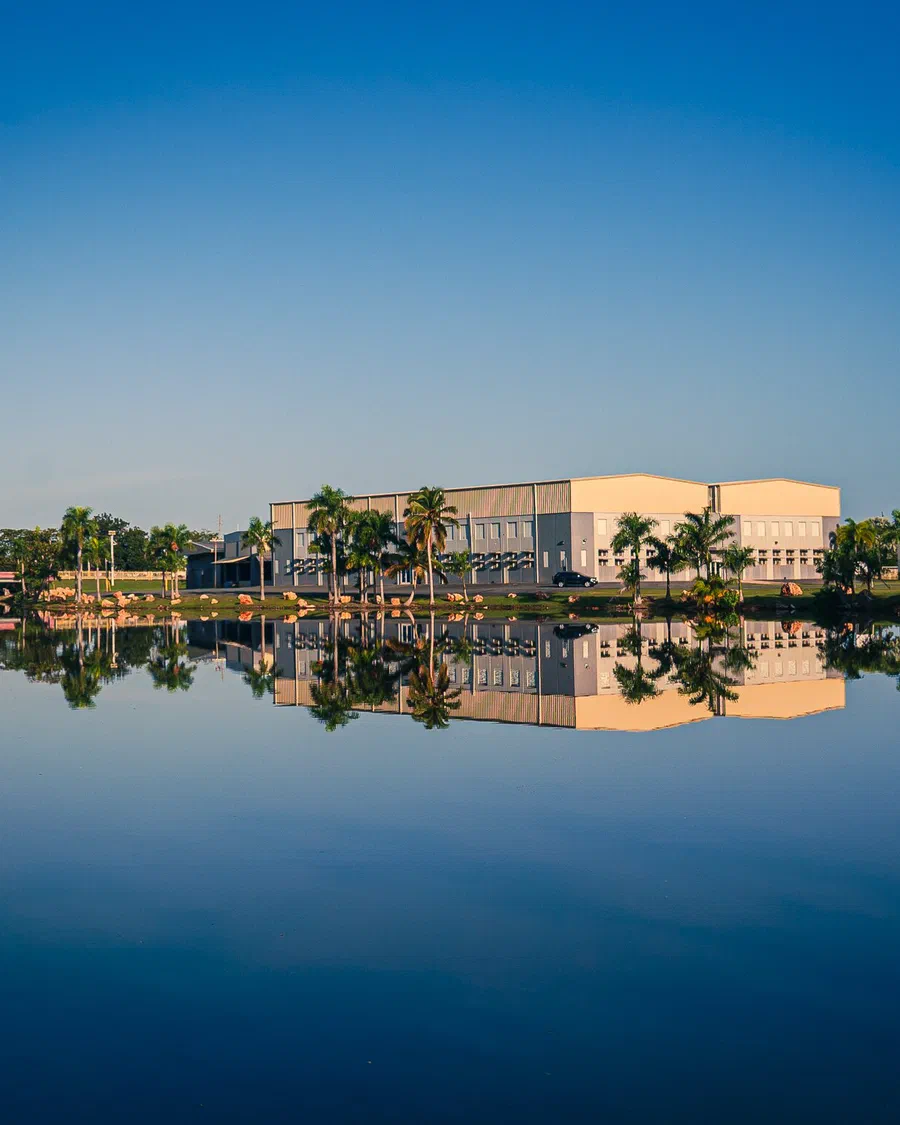 Un edificio claro entre palmas se refleja completo en el agua quieta bajo un cielo azul, con un puente rosado a la izquierda, en Levittown, Puerto Rico